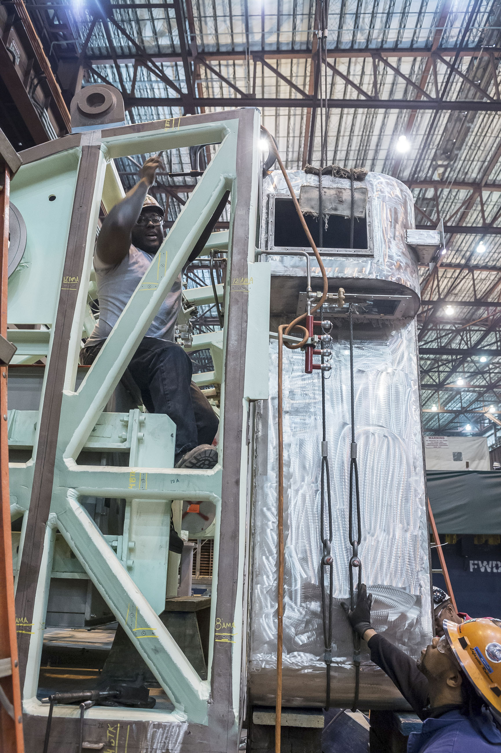 Sheet metal worker Ricky Campbell clamps a charcoal filter in place as crane operator Lafayette Roberson in orange hardhat fites the filter  to the fan room for the Virginia-class submarine Indiana (SSN 789).
